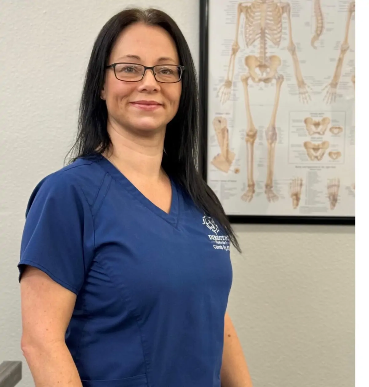 Cassie Fry, PTA, in navy scrubs in a treatment room at Direct PT of Huntsville, beside an anatomy chart.