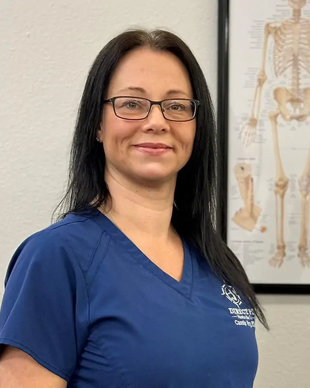 Cassie Fry, PTA, founder of Direct PT of Huntsville, in navy clinic scrubs beside an anatomy chart.